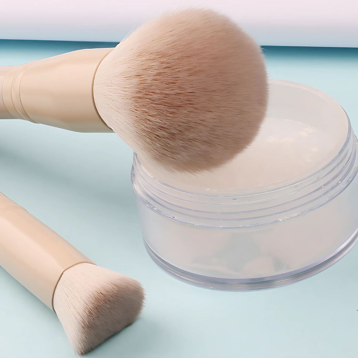 Zwei Make-up-Pinsel mit hellbeigen Griffen neben einem durchsichtigen Behälter mit weißem Puder.
