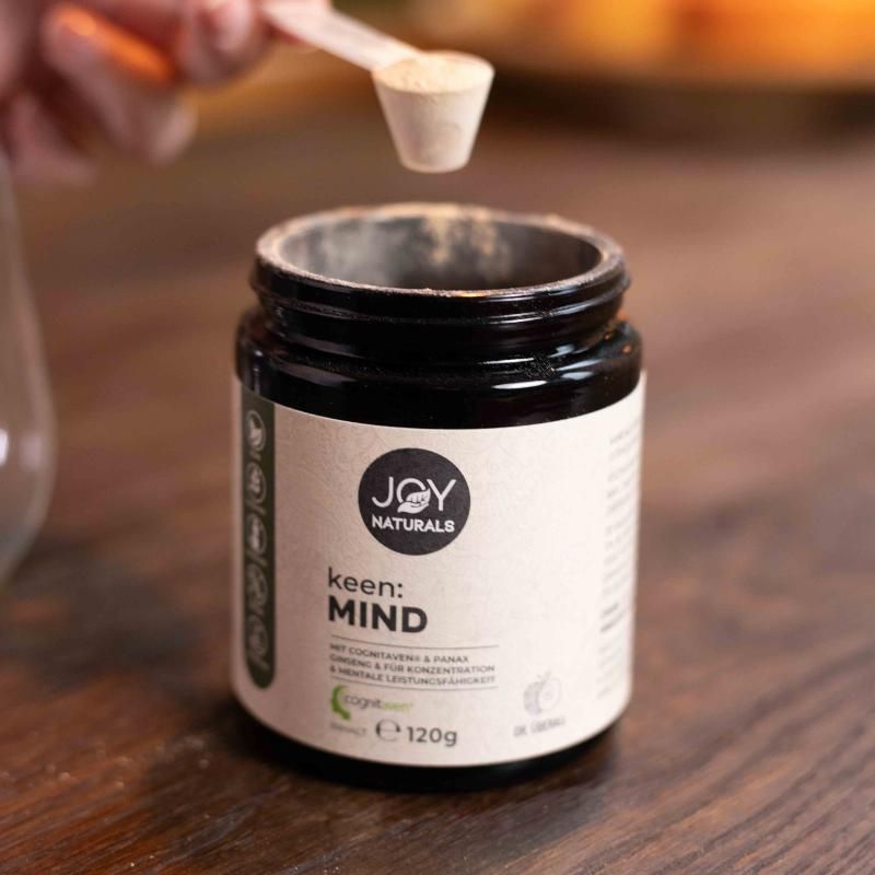 Braunes Glas mit JOY NATURALS keen:MIND Pulver. Ein Messlöffel wird in das Glas gehalten. Aufschrift: 120g.