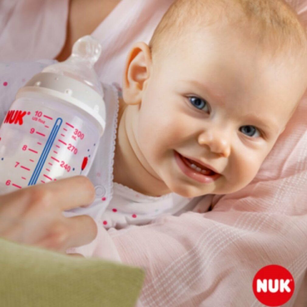 Baby hält Babyflasche. Aufdrucke: NUK, Messskala. Baby lächelt.