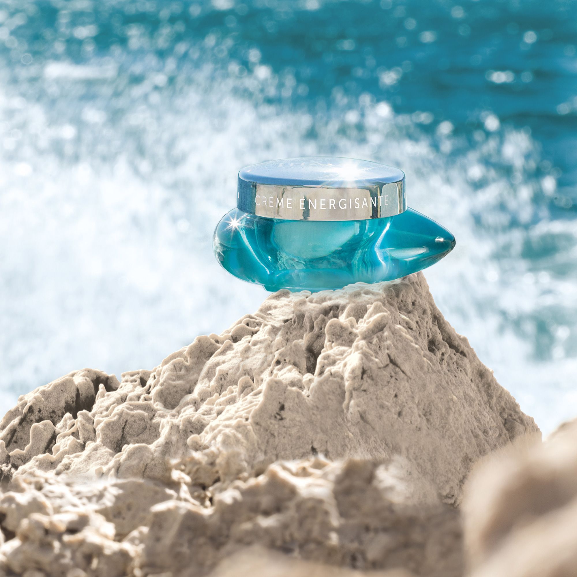 Türkisfarbener Tiegel mit silbernem Deckel auf einem Felsen am Meer. Auf dem Deckel steht "CRÈME ÉNERGISANTE".