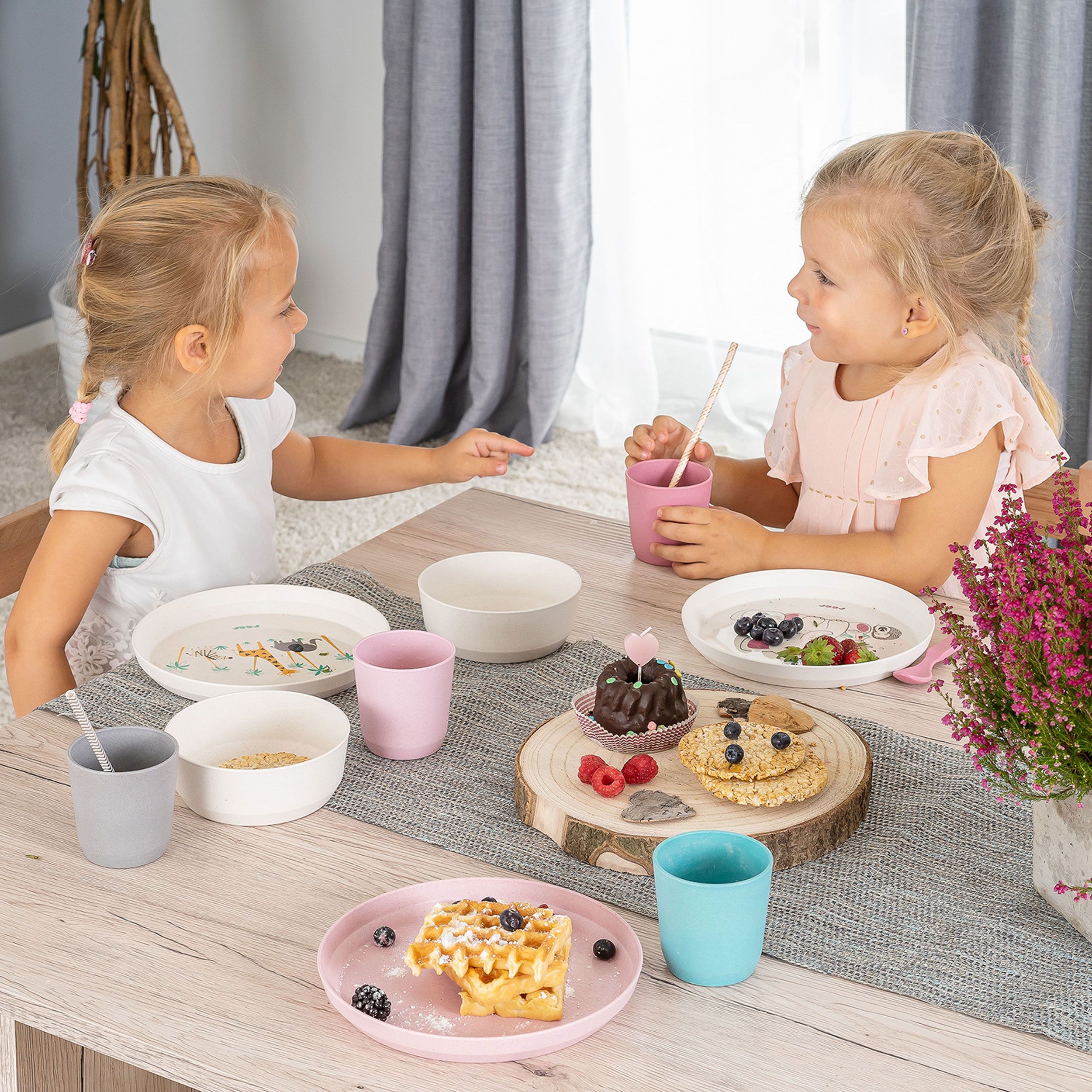 Kindergeschirr-Set mit Tellern, Schalen und Bechern. Auf den Tellern sind Tiermotive abgebildet. Kinder sitzen am Tisch.