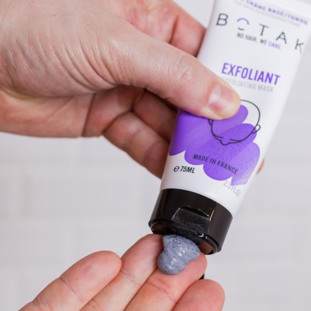 Hand drückt graues Peeling aus einer Tube. Tube: BOTAK, EXFOLIANT, 75ml. Hintergrund: weiße Fliesen.