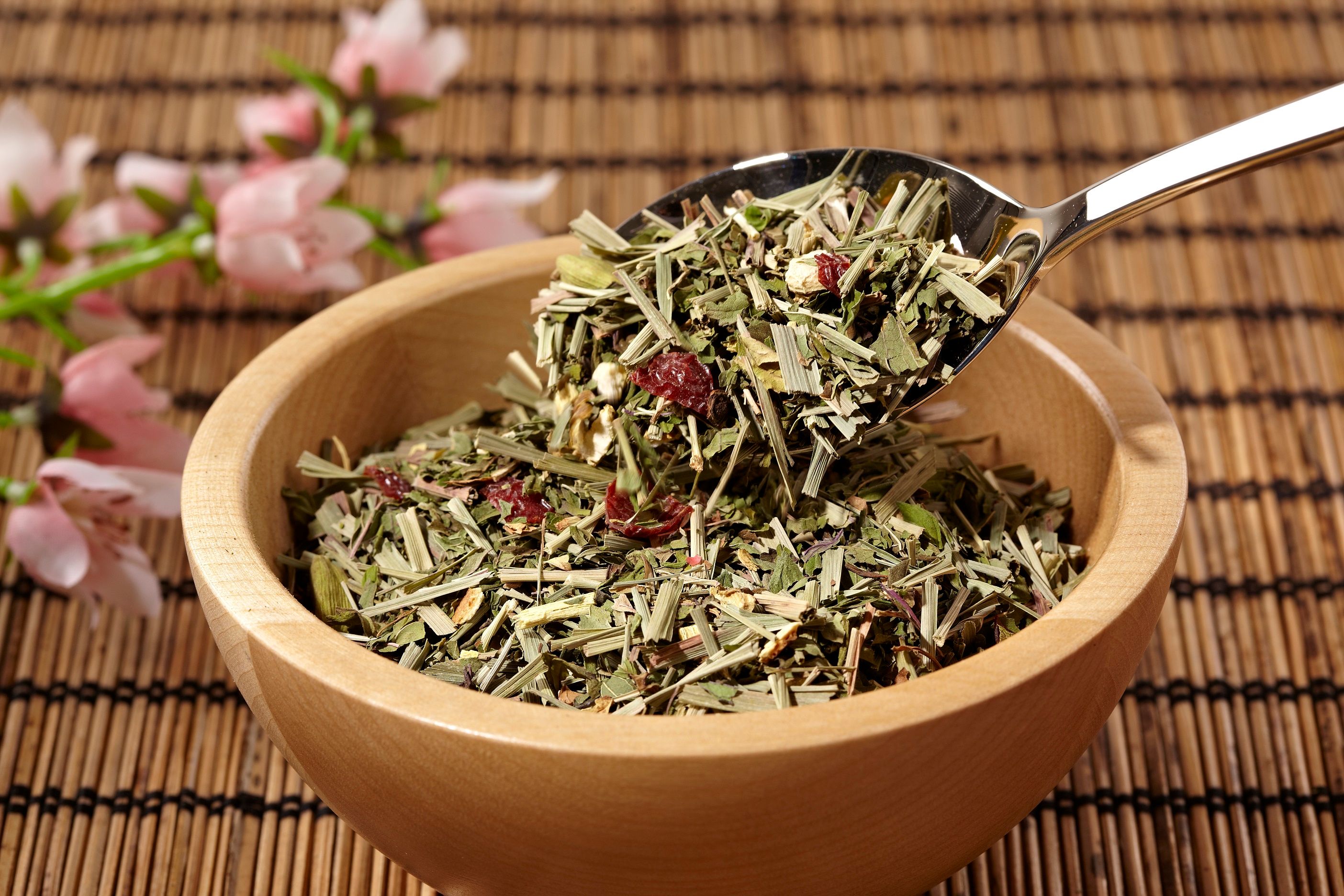 Holzschale mit Kräutermischung. Ein Löffel hebt Kräuter aus der Schale. Im Hintergrund Blüten und Bambusmatte.