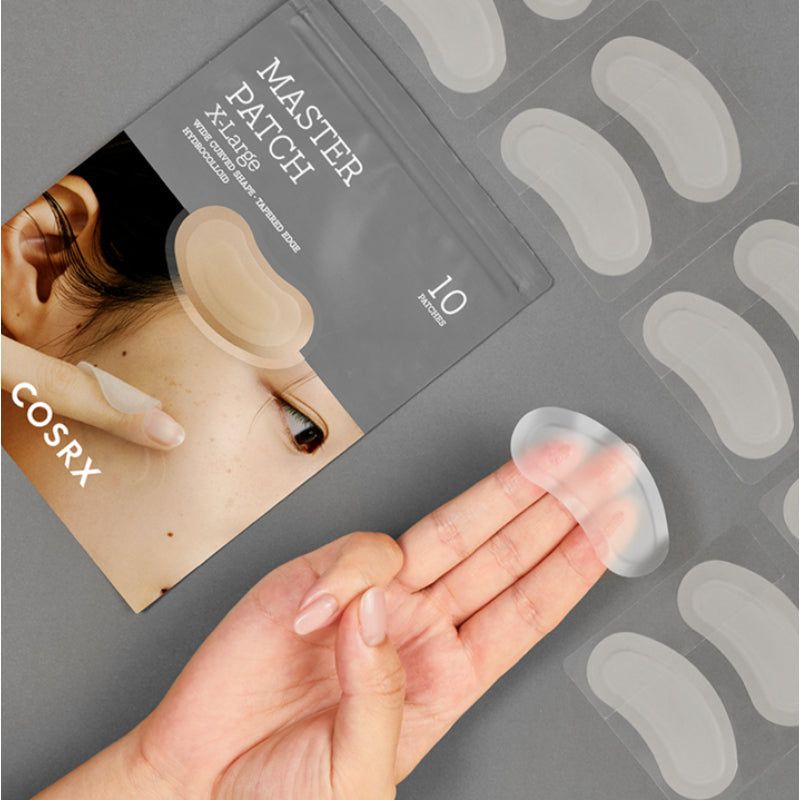 Eine Hand hält ein transparentes, ovales Pflaster. Im Hintergrund sind weitere Pflaster und eine Verpackung zu sehen.