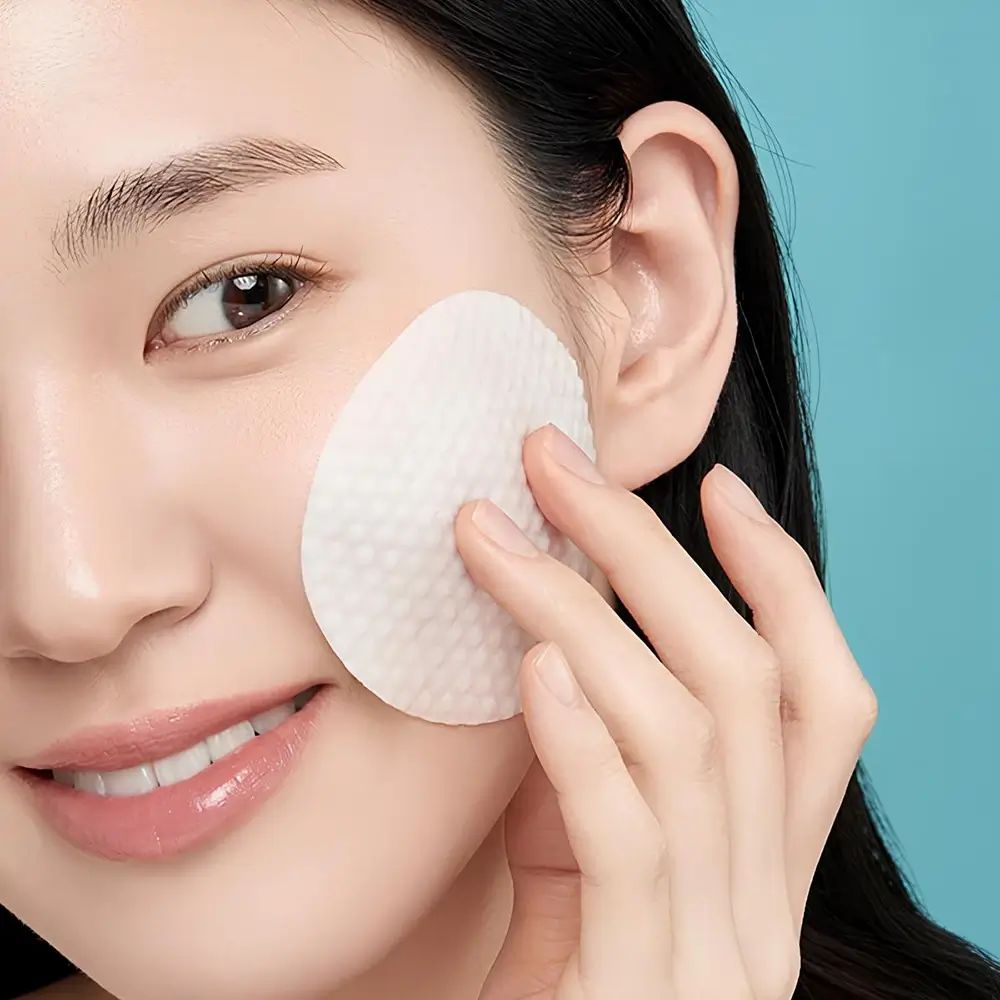 Frau mit einem Pad auf der Wange. Das Pad ist weiß und hat eine strukturierte Oberfläche. Blauer Hintergrund.