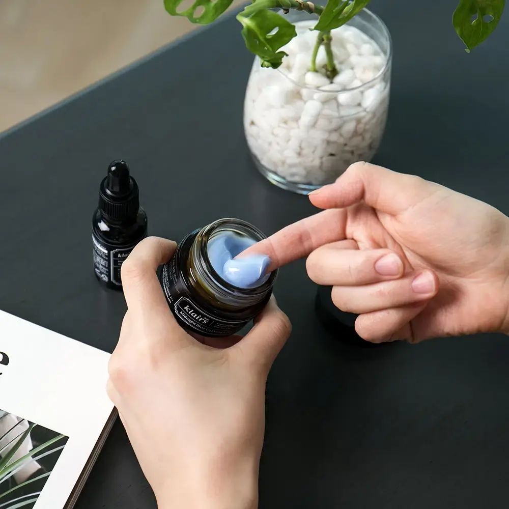 Hände halten ein offenes Glas mit blauer Creme. Daneben eine Flasche mit schwarzem Etikett.