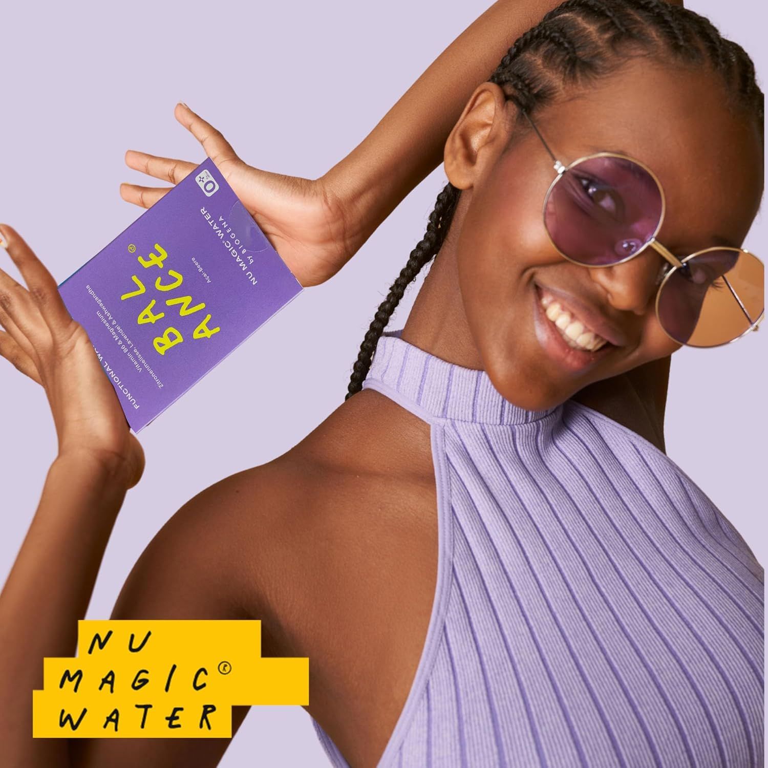 Frau hält lila Schachtel mit "BALANCE" und "NU MAGIC WATER" von BIOGENA. Sie trägt Sonnenbrille und lila Top. Produkt im Fokus.