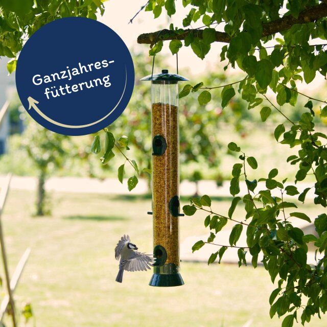 Vogelfutterspender aus Kunststoff, gefüllt mit Streufutter. Beschriftung: Ganzjahresfütterung. Vogel beim Fressen.