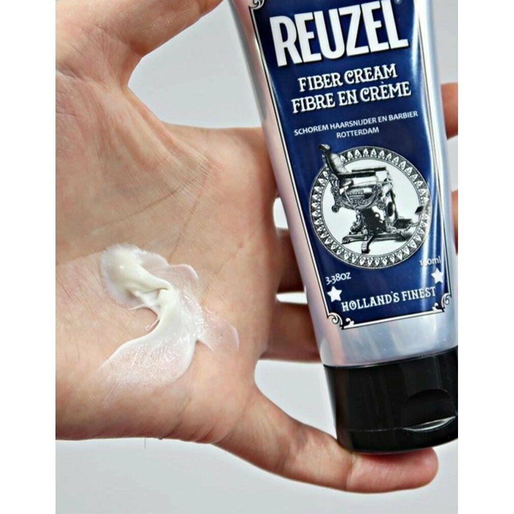 Hand mit Creme. Tube mit blauem Etikett. Aufschrift REUZEL FIBER CREAM. Abbildung eines Friseurstuhls.