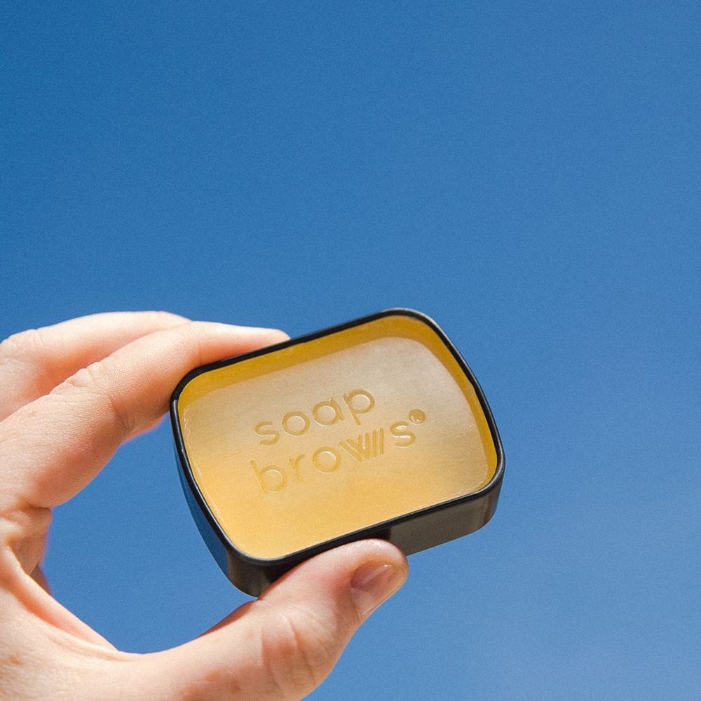Hand hält eine offene, schwarze Dose mit gelbem Inhalt. Aufschrift 'soap brows'.