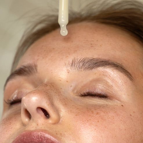 Gesicht einer Person mit geschlossenen Augen. Eine Pipette tropft eine Flüssigkeit auf die Stirn. Haut mit Sommersprossen.