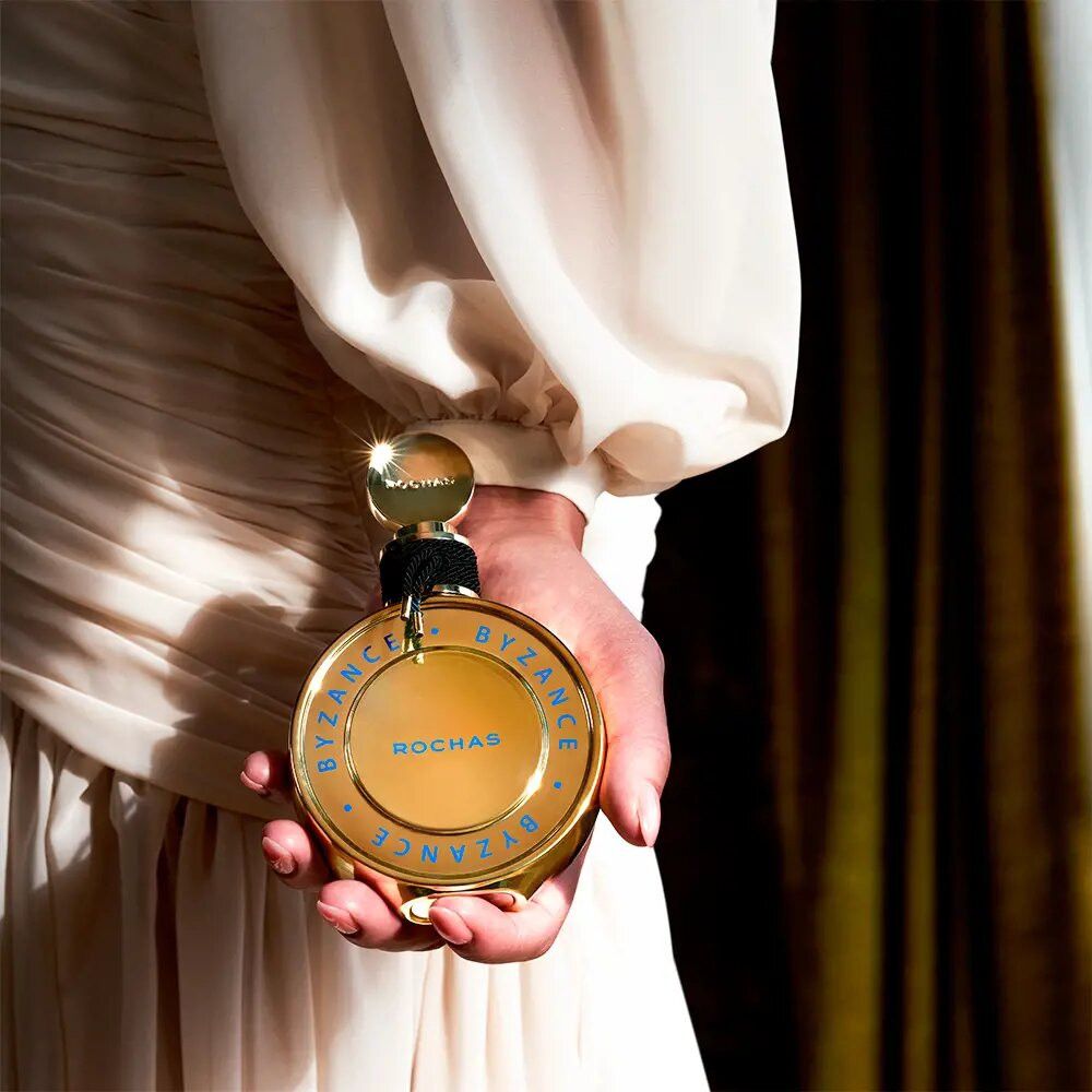Goldfarbener Flakon, gehalten von einer Person. Aufschrift: Byzance, Rochas. Schwarze Kordel am Hals.