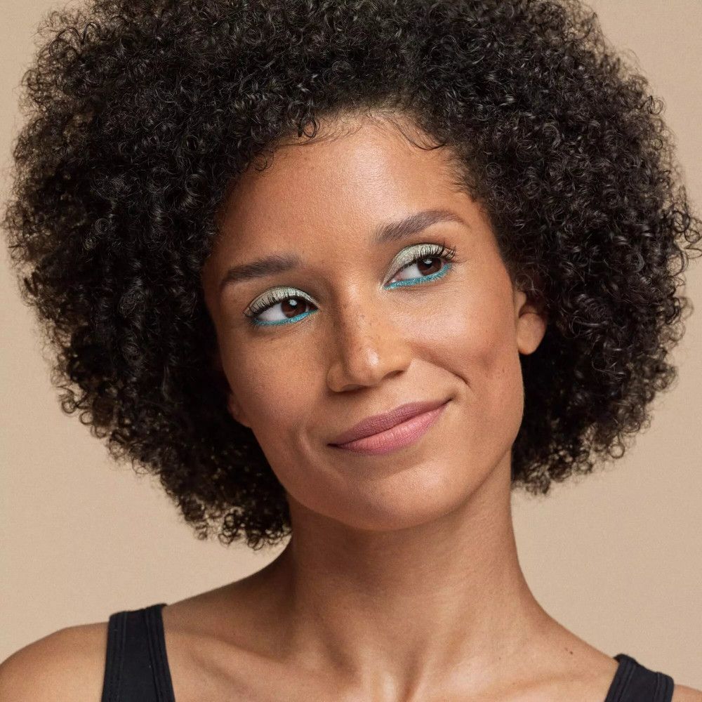 Porträt einer Frau mit blauem Eyeliner und rosafarbenem Lippenstift. Sie lächelt und trägt ein schwarzes Top.