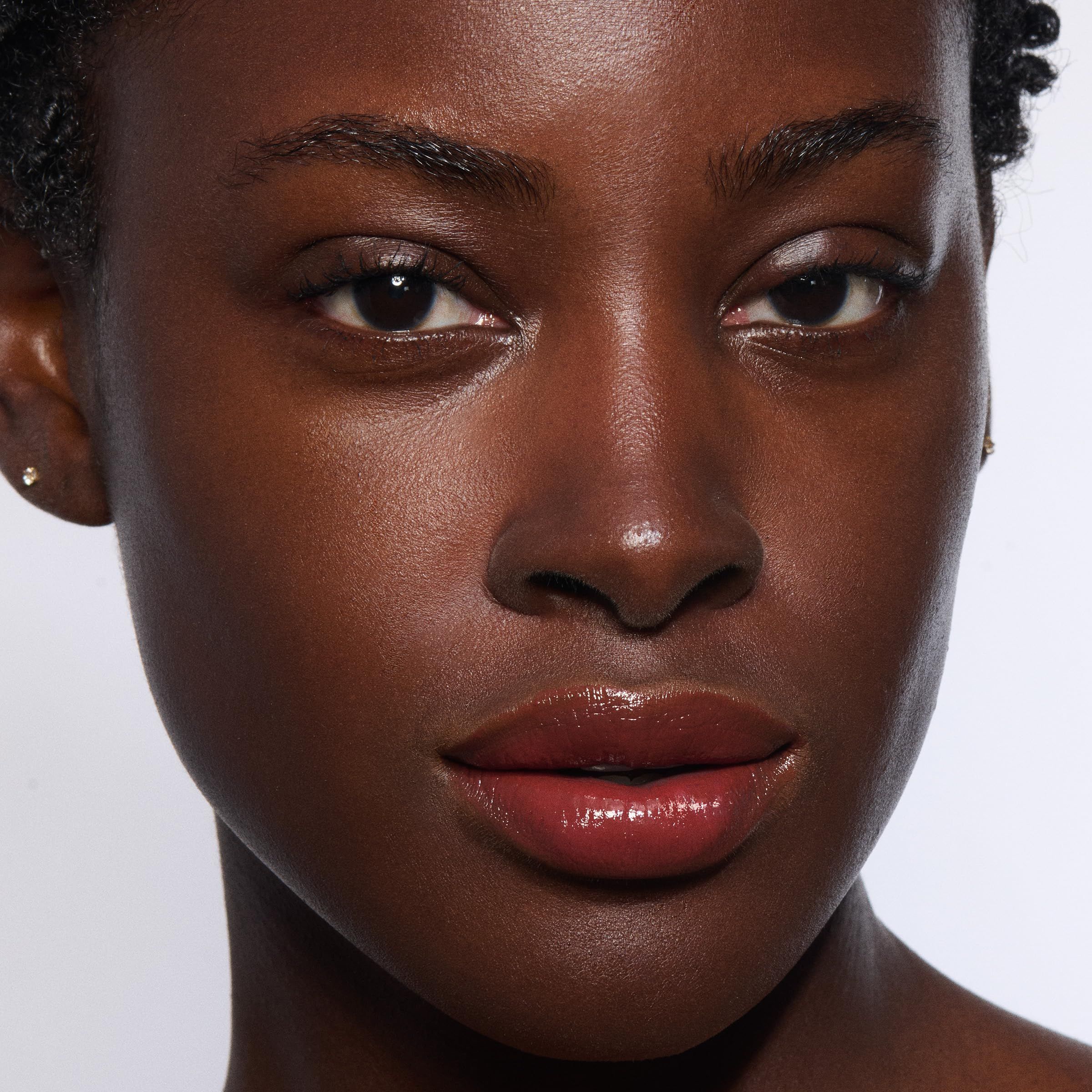 Nahaufnahme eines Gesichts mit Lippenstift. Dunkle Haut, Lippen mit Lip Oil Balm. Keine Produktnamen sichtbar.