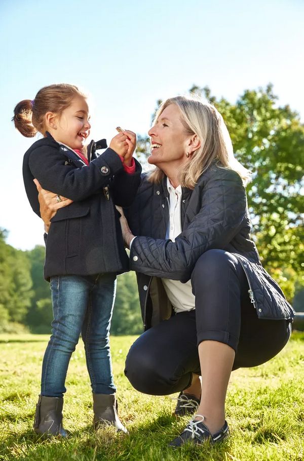 Frau und Kind im Freien. Frau kniet, Kind steht. Beide lächeln. Frau trägt dunkle Jacke, Kind blaue Jacke.