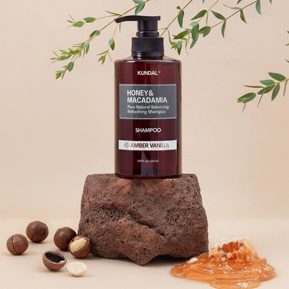 Shampoo-Flasche auf Stein. Text: KUNDAL, HONEY & MACADAMIA, SHAMPOO, AMBER VANILLA. Honigwabe, Macadamia-Nüsse, grüne Blätter.