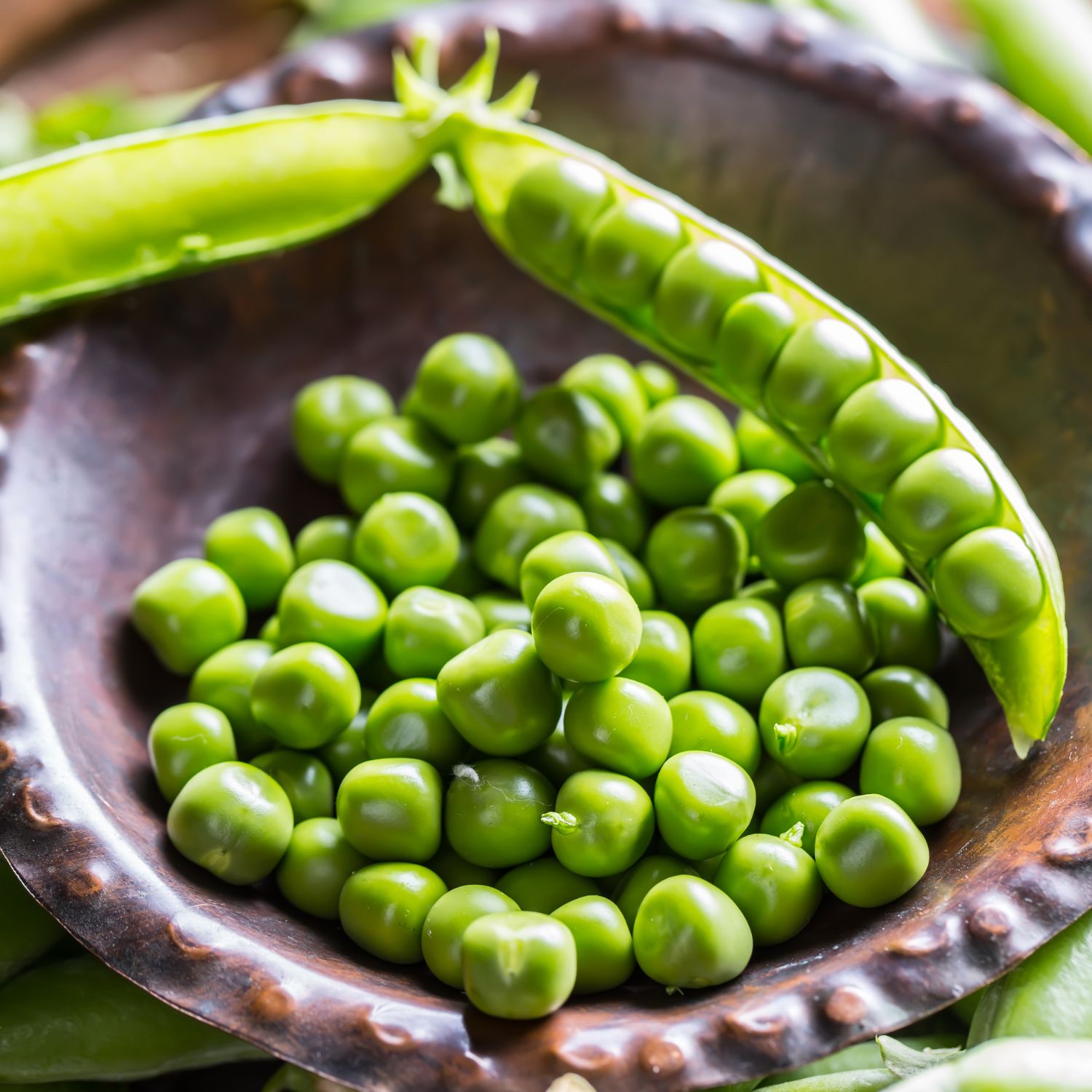 Braune Schale mit grünen Erbsen und einer geöffneten Erbsenschote. Die Erbsen sind hellgrün. Die Schale hat eine unebene Oberfläche.