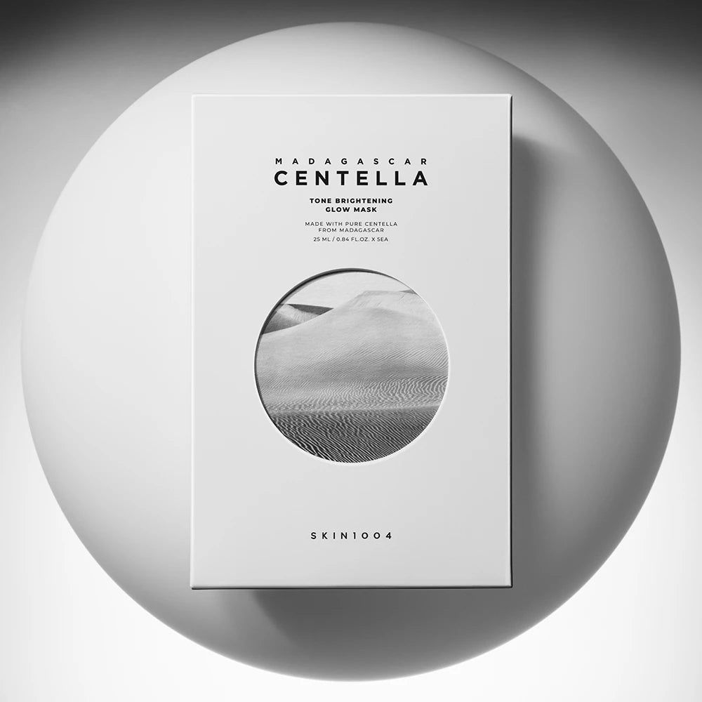 Weiße Produktverpackung mit Text und rundem Bild. Aufschrift: MADAGASCAR CENTELLA Tone Brightening Glow Mask. Marke: SKIN1004. Auf weißem Hintergrund.