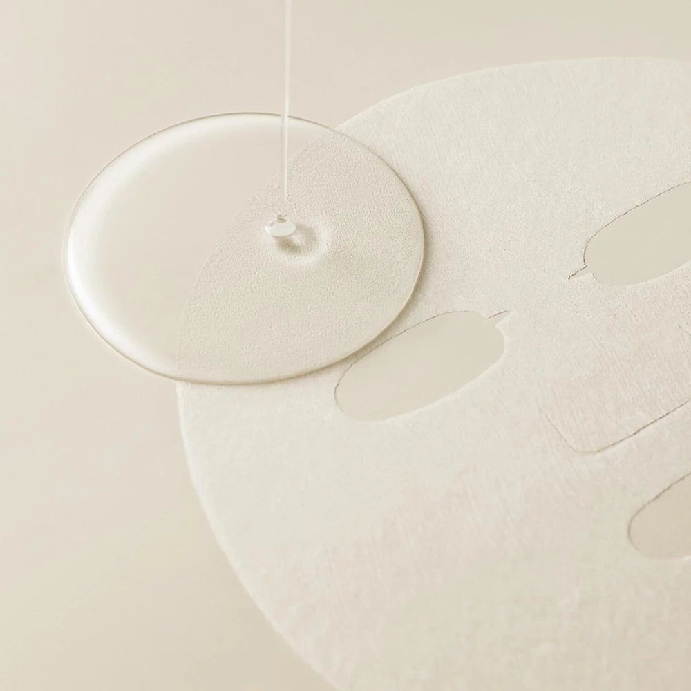 Klare Flüssigkeit tropft auf eine runde Maske. Maske mit Aussparungen für Augen und Mund. Hintergrund hell.