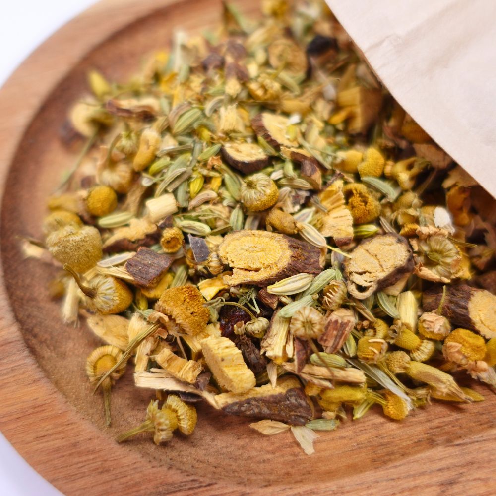 Getrocknete Kräuter und Blüten in einer hölzernen Schale, teilweise von einer Papiertüte verdeckt. Enthält Kamille, Fenchel und Wurzeln.