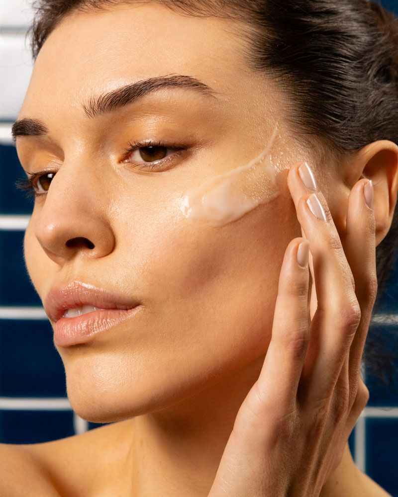 Frau trägt Creme auf ihr Gesicht auf. Helle Haut, Produkt auf der Wange. Blauer Hintergrund.