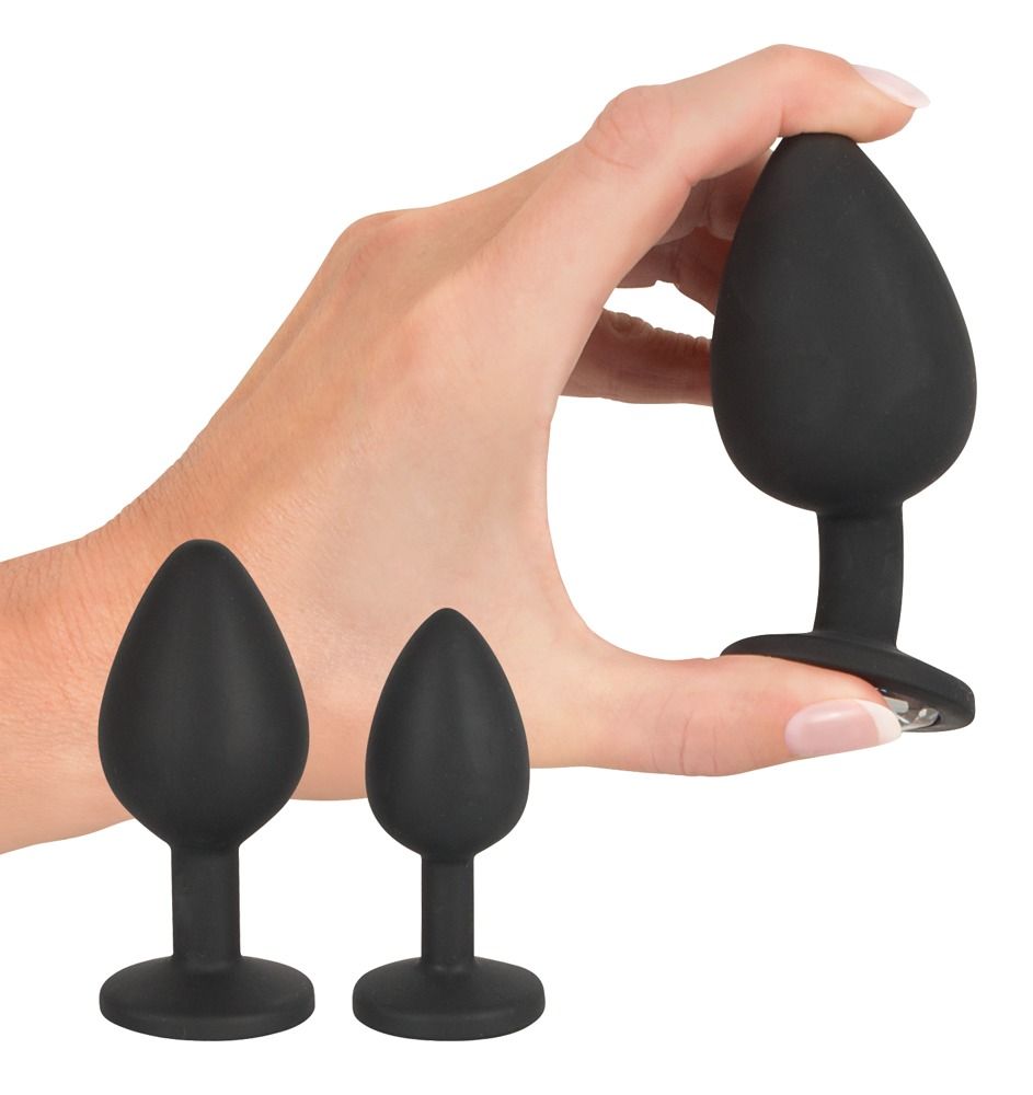 Hand hält einen schwarzen Anal-Plug. Zwei weitere Plugs liegen daneben. Alle aus Silikon.