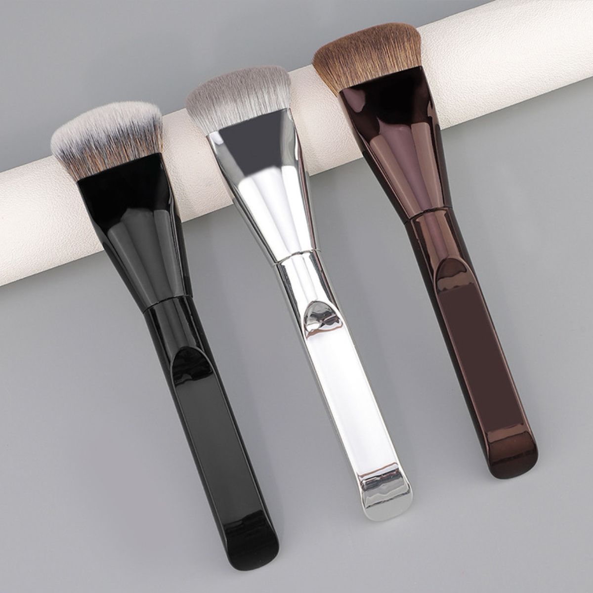 Drei flache Make-up-Pinsel mit schwarzen, silbernen und braunen Griffen und dichten Borsten.