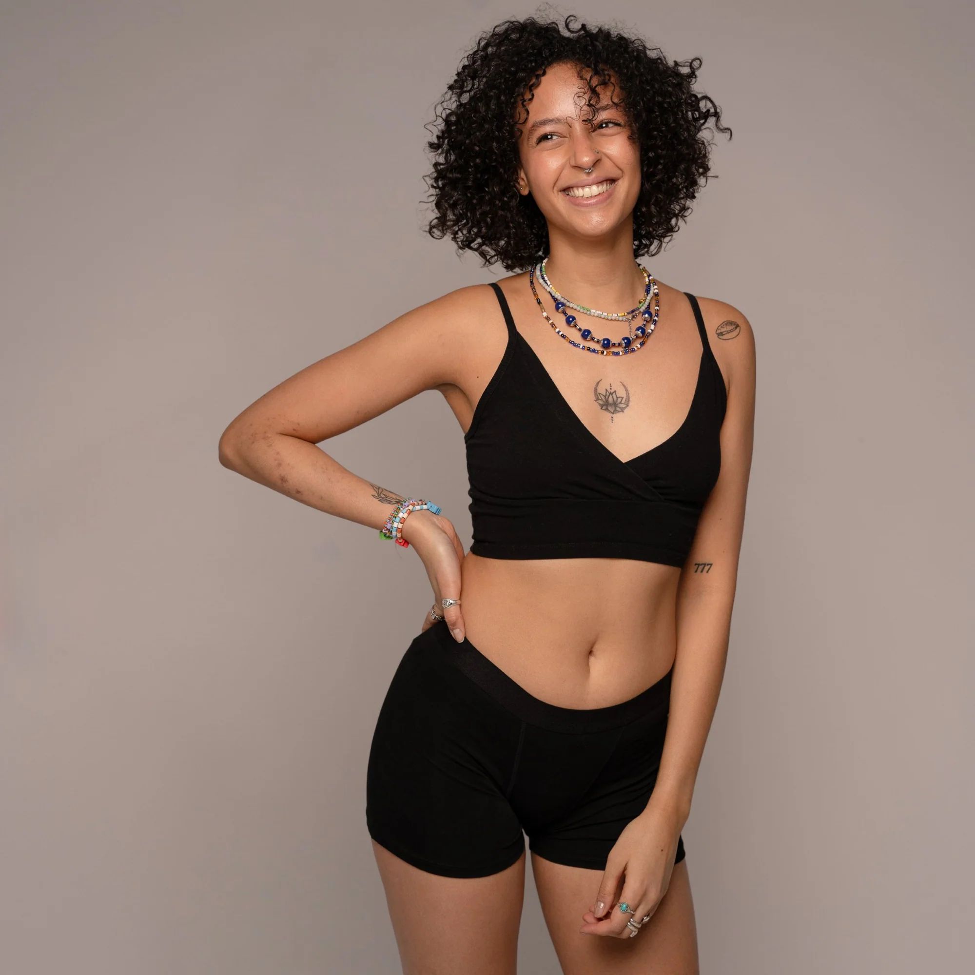 Person trägt schwarze Unterhose und ein schwarzes Top. Person lächelt. Mehrere Halsketten und Armbänder.