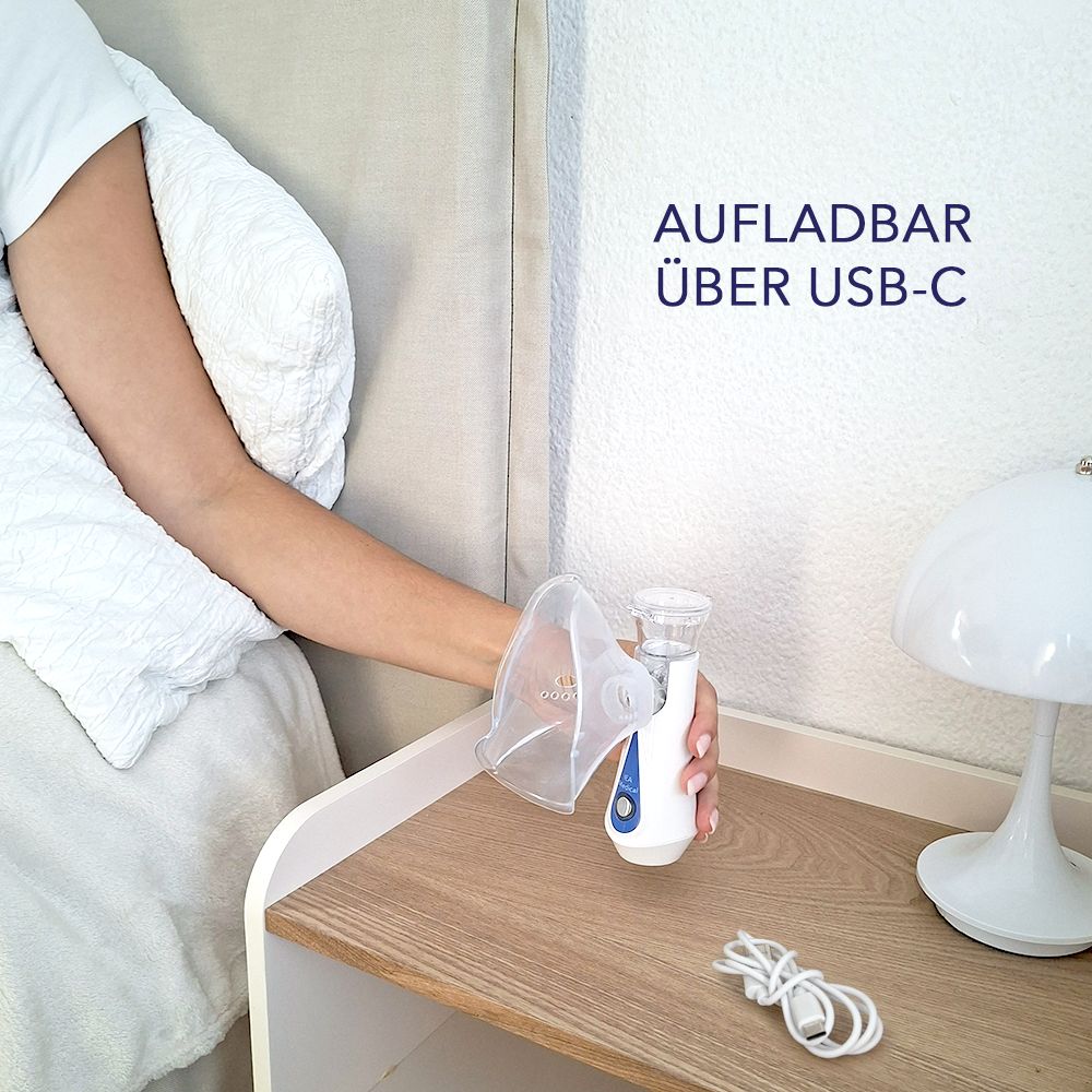 Inhalationsgerät mit Maske auf Nachttisch. Gerät weiß-blau, Maske transparent. USB-C Kabel liegt daneben. Text: Aufladbar über USB-C.