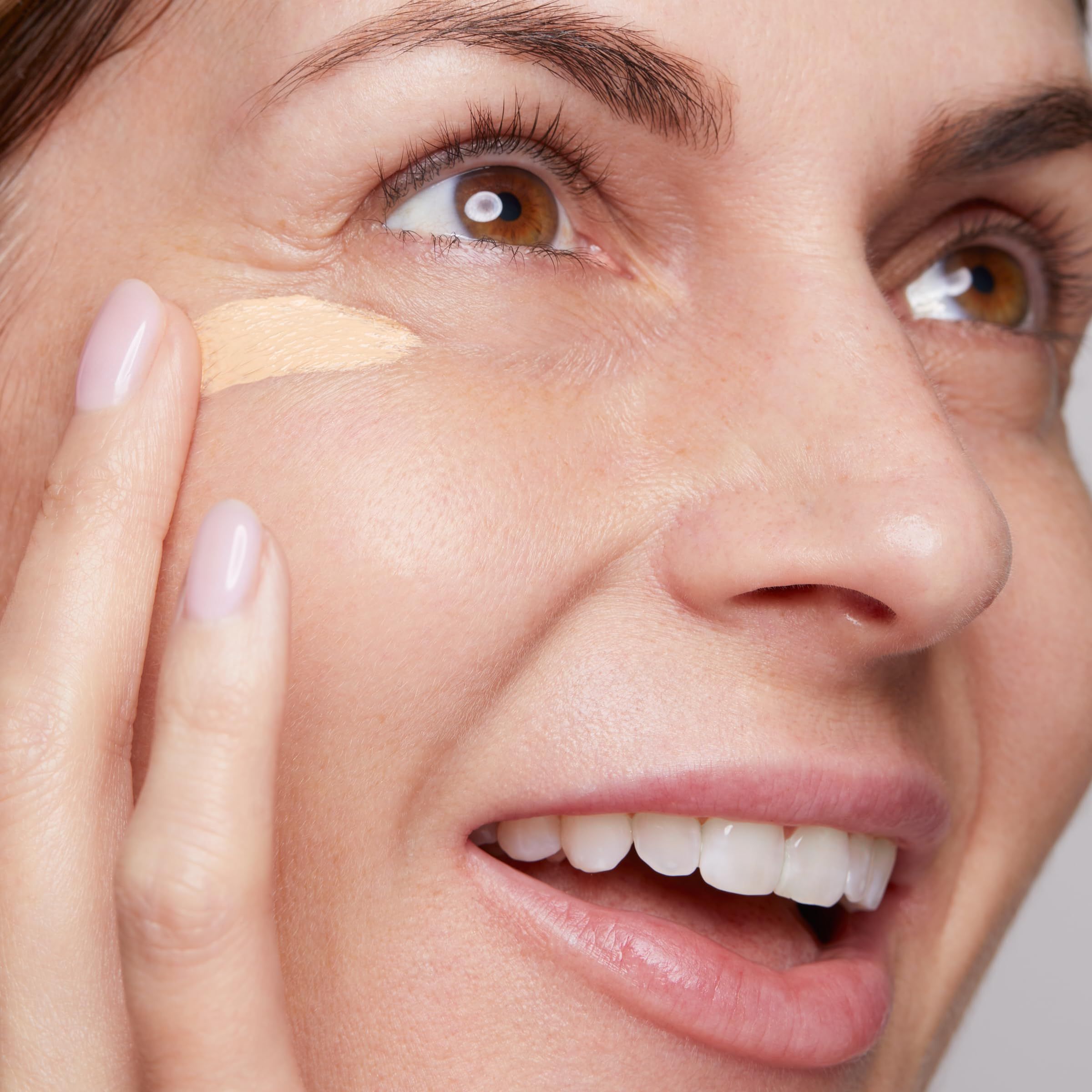 Frau trägt Produkt auf. Beige Produkt auf der Haut. Gesichtsausdruck: lächelnd.