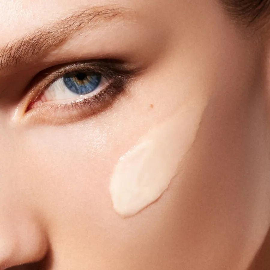 Nahaufnahme eines Auges und einer Hautpartie mit aufgetragener Creme. Creme-Farbe: hellbeige.