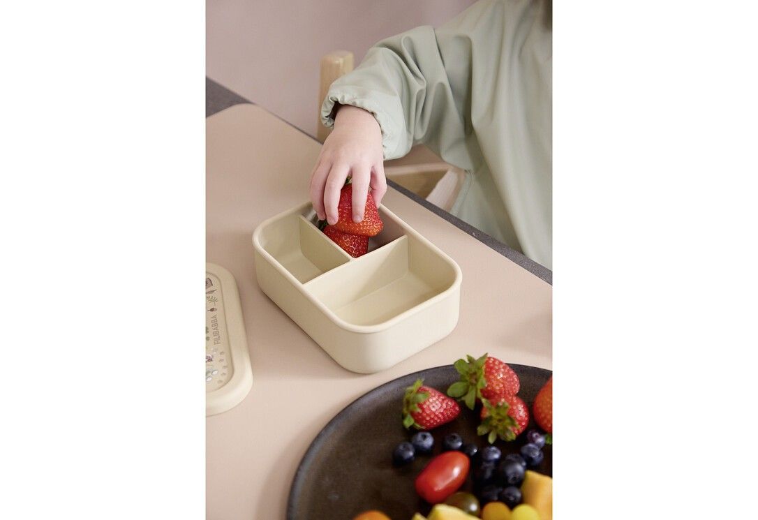 Kind greift in beige Brotdose mit Erdbeeren. Deckel daneben. Aufteilung in drei Fächer. Auf Tisch mit Obst.