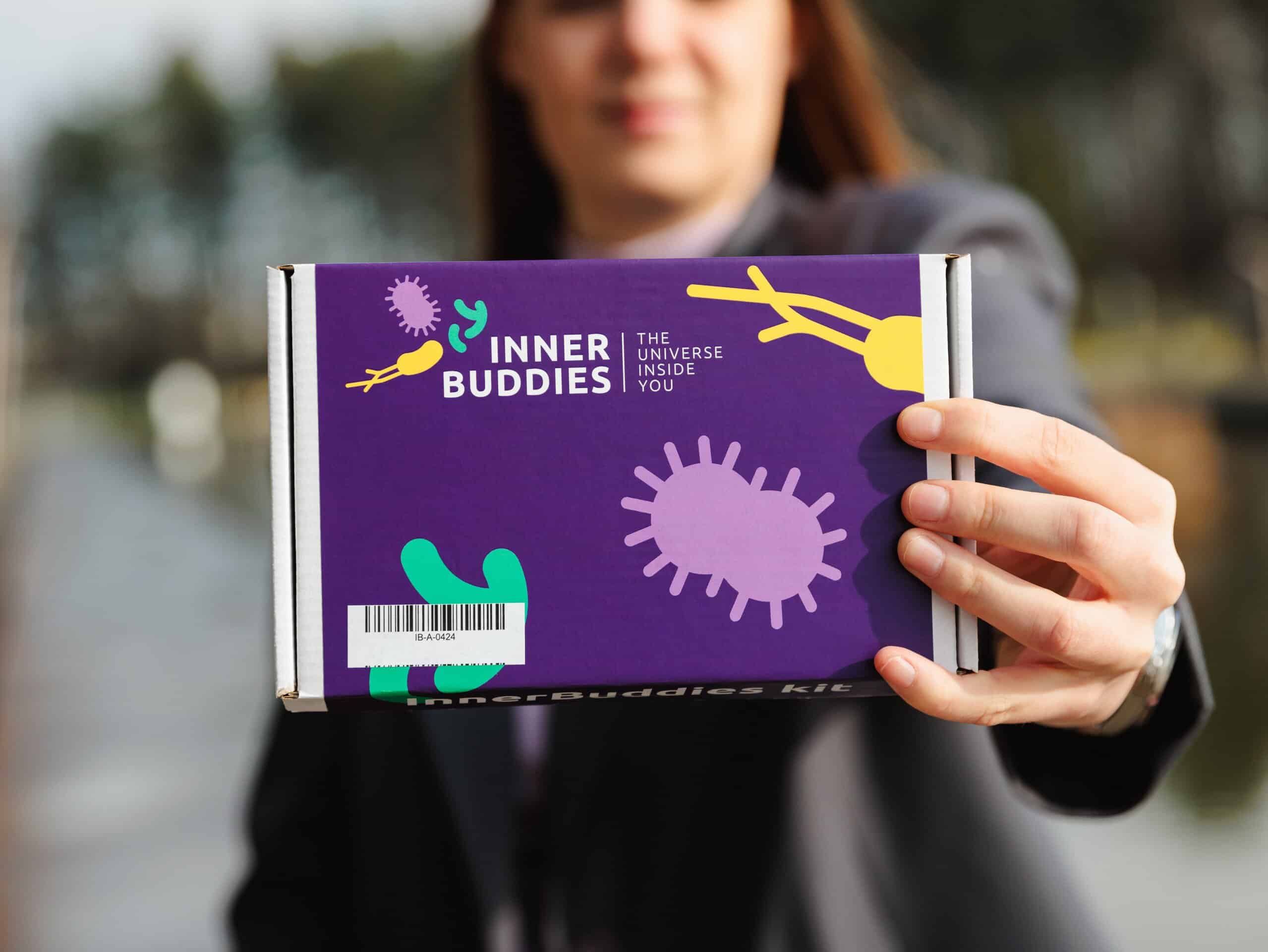 Person präsentiert eine Inner Buddies Box mit lila Hintergrund und bunten Mikroben-Illustrationen.