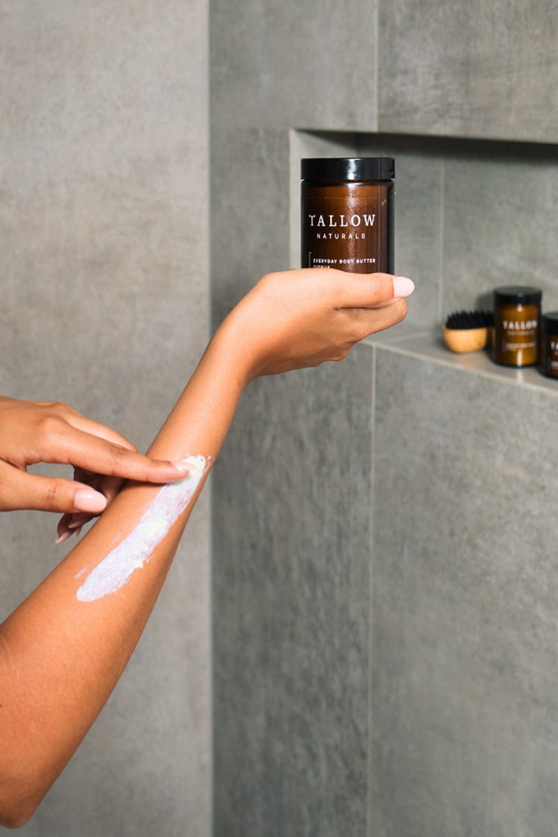 Person trägt Körperbutter auf Arm auf. Dose mit Aufschrift: TALLOW NATURALS, EVERYDAY BODY BUTTER. Grauer Hintergrund.
