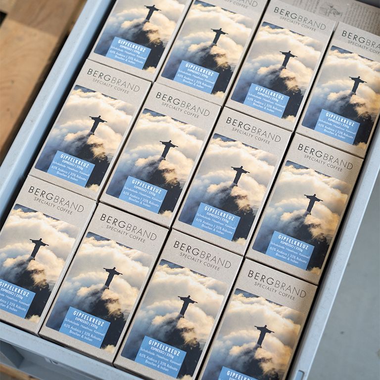 Mehrere Kaffeepackungen in einer grauen Kiste. Auf jeder Packung steht BERGBRAND Gipfelkreuz Specialty Coffee.