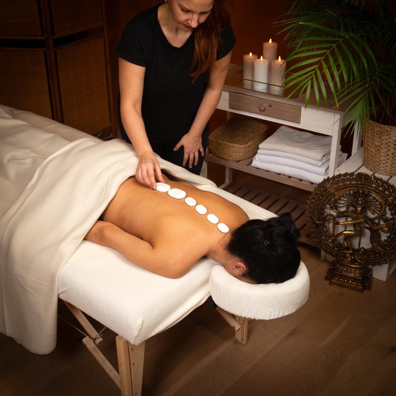 Person legt weiße, ovale Steine auf den Rücken einer Person. Auf einer Liege.