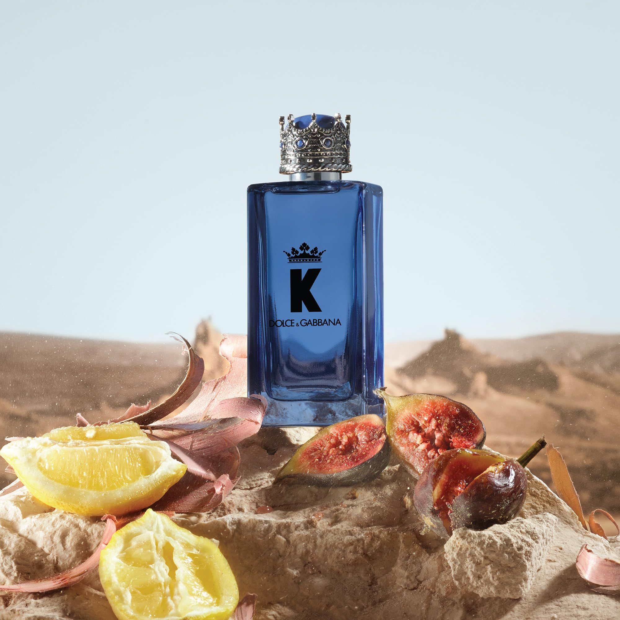 Blaues Parfümflakon mit Krone. Aufschrift "K" und "Dolce & Gabbana". Zitronen und Feigen auf Stein. Wüstenhintergrund.