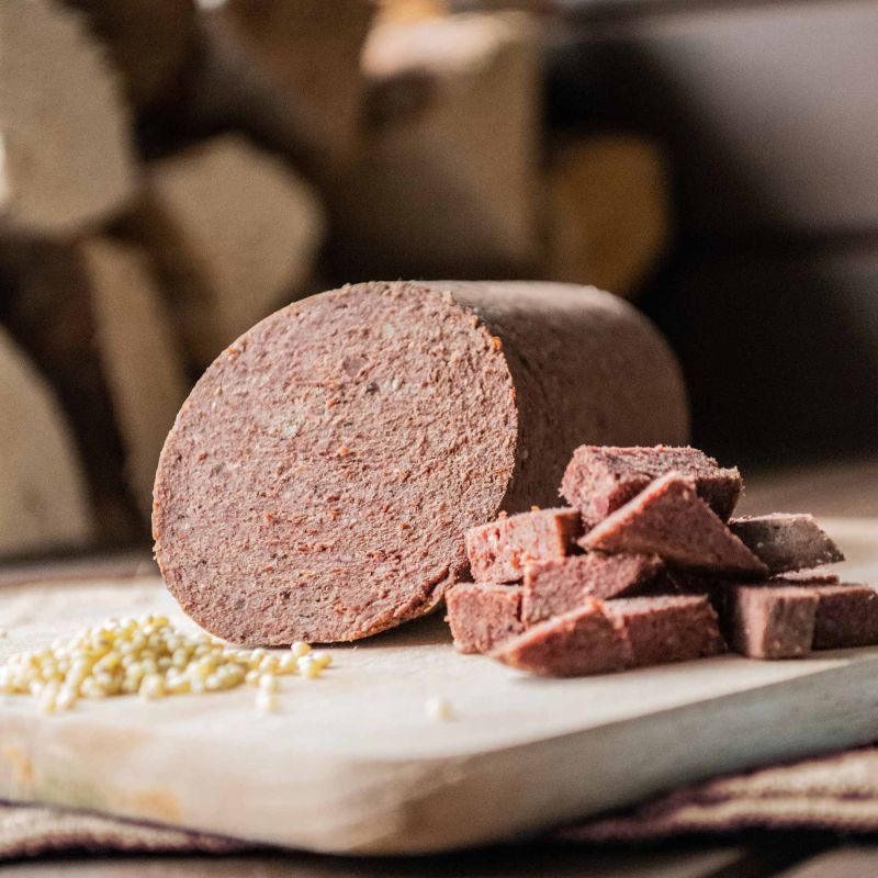 Aufgeschnittene Wurst, Würfel und Körner auf Holzbrett. Fleischfarben. Im Hintergrund Holzstücke.