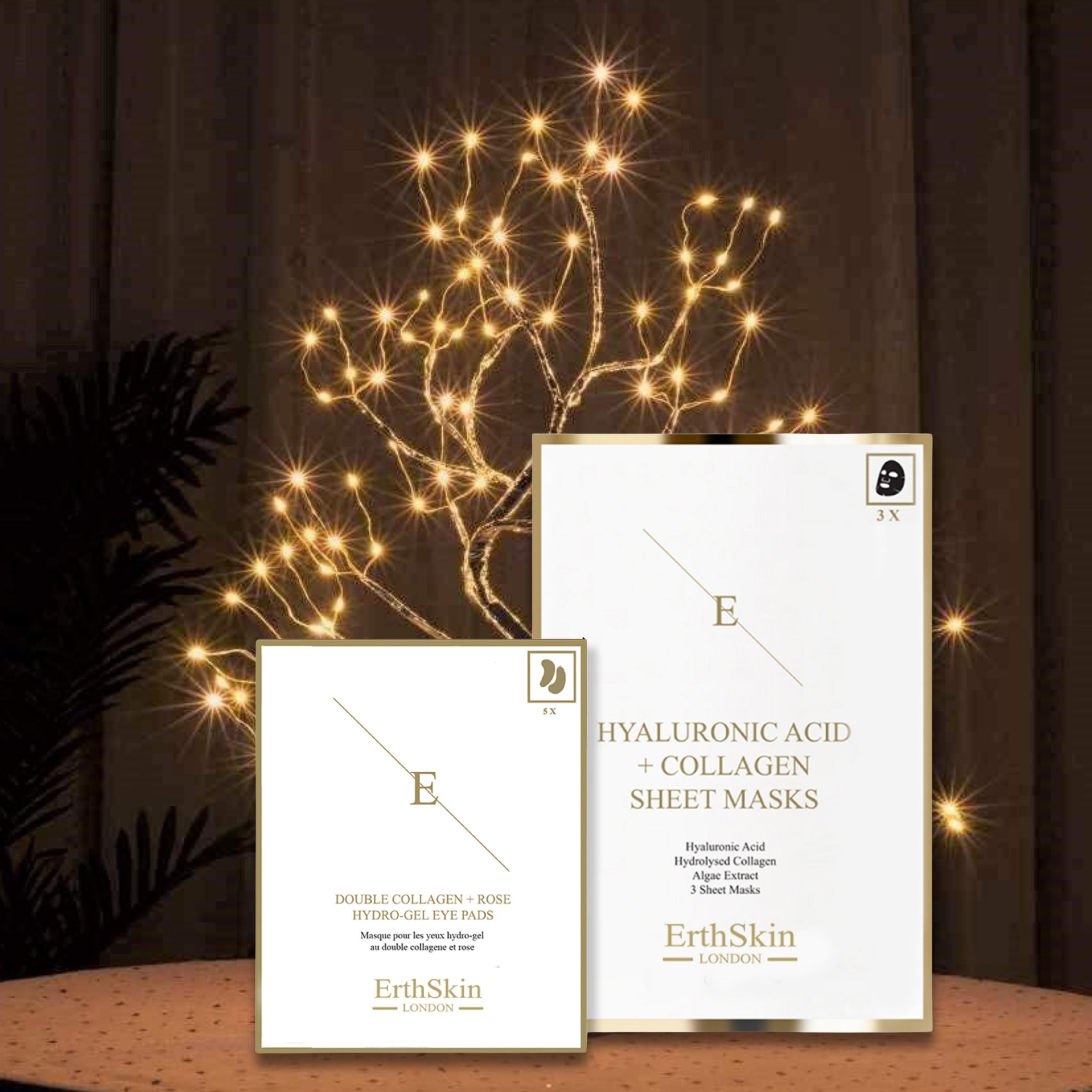 Zwei weiße Produktverpackungen mit goldfarbenen Akzenten. Eine Packung mit Augenpads, die andere mit Gesichtsmasken. Im Hintergrund eine dekorative Lichterkette.