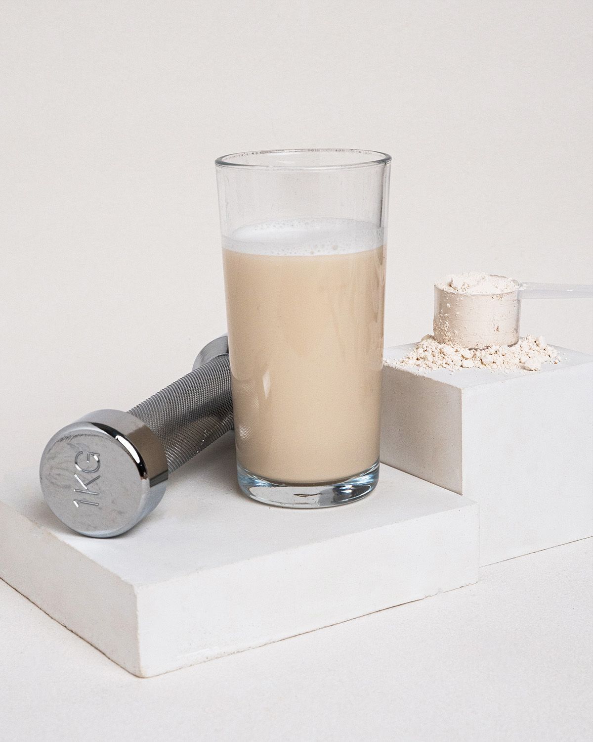 Glas mit Proteinshake, Hantel und Messlöffel mit Pulver auf weißen Blöcken. Der Shake ist hellbraun.