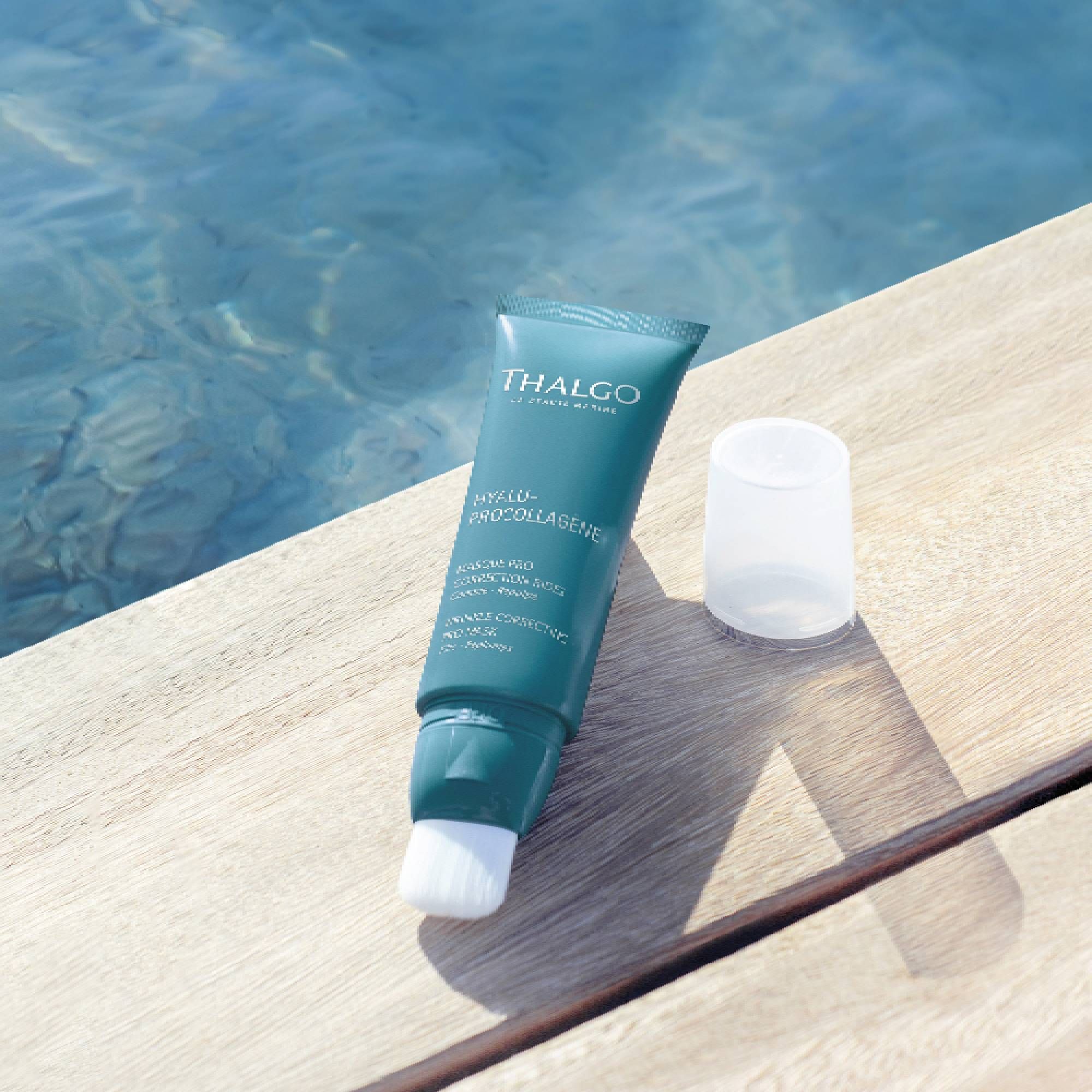 Türkisfarbene Tube mit weißem Pinsel und abgenommener Kappe. THALGO-Logo und Produktbezeichnung. Auf Holzoberfläche.
