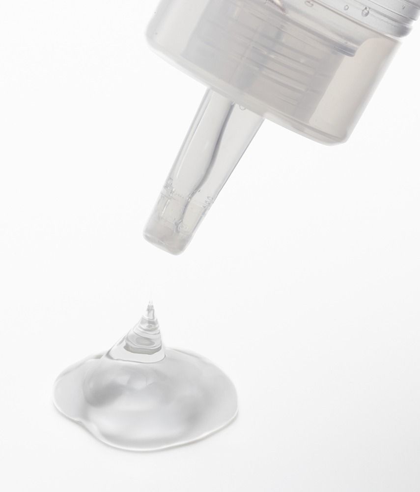 Aus einer Flasche tropft transparente Flüssigkeit. Flüssigkeit bildet einen Tropfen auf weißem Hintergrund.