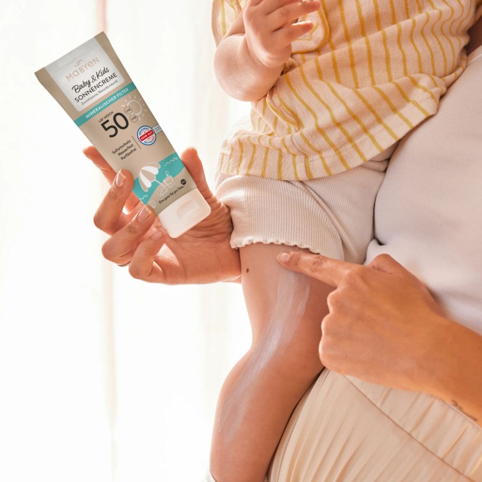Person hält Sonnencreme-Tube. Marke: MABYEN Baby & Kids. LSF 50. Creme wird auf Bein aufgetragen. Kind im Hintergrund.