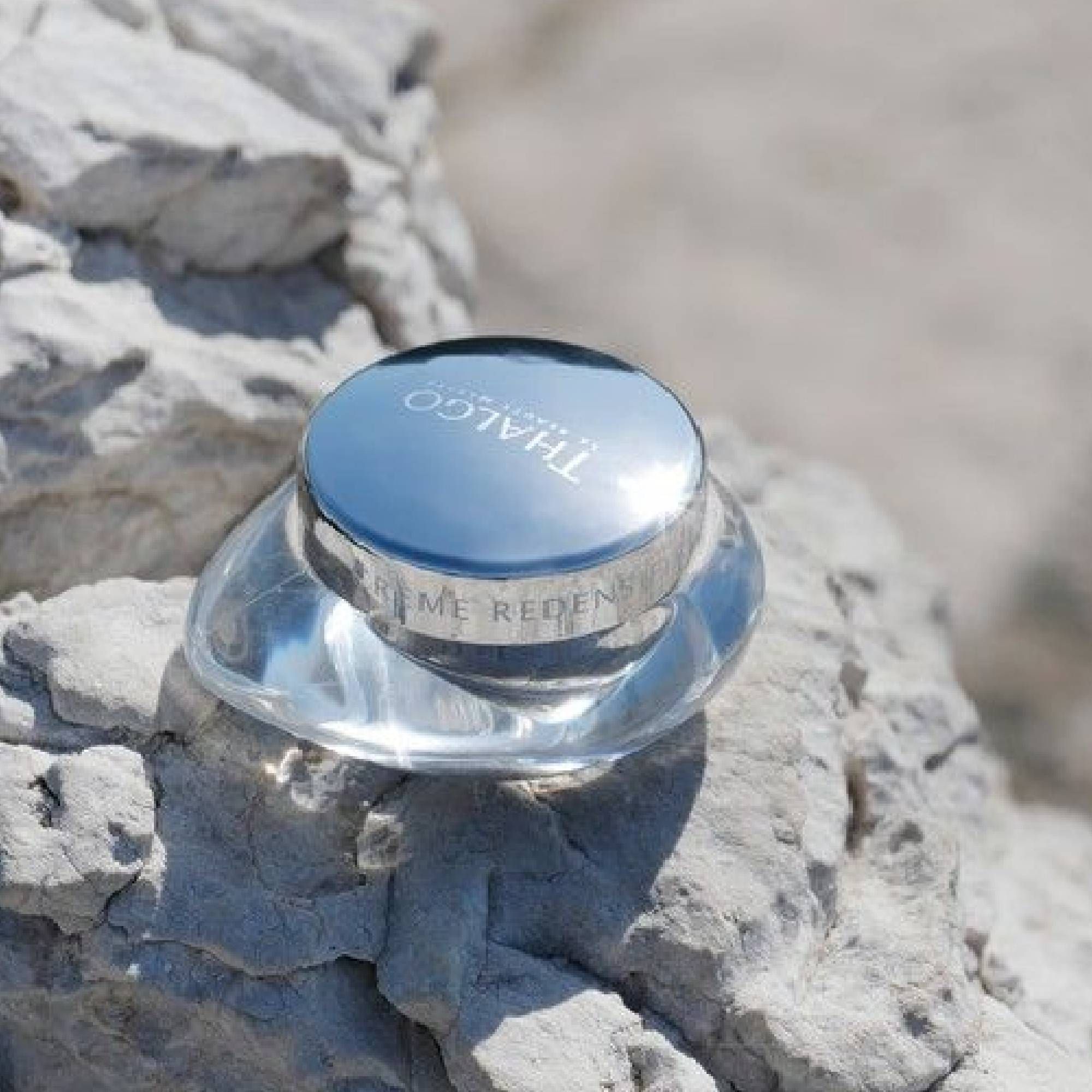 Glasbehälter mit silbernem Deckel auf Felsen. Aufschrift: THALGO, Crème Redensifiante. Klare, organische Form.