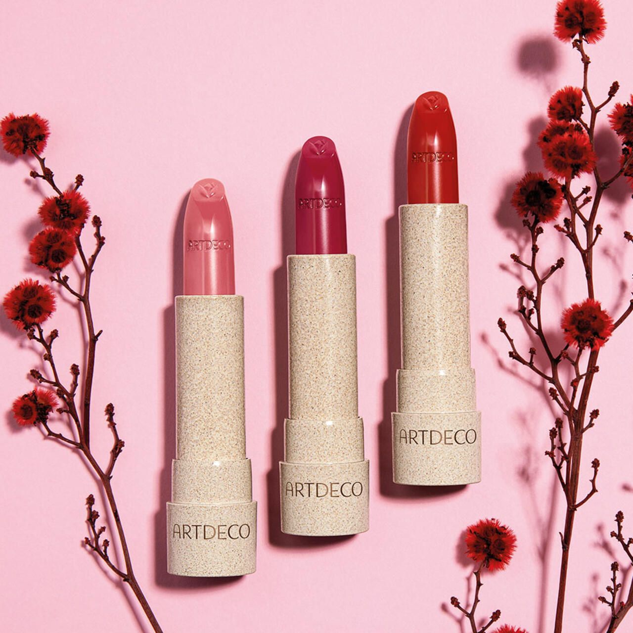 Drei Lippenstifte in verschiedenen Rottönen. Cremefarbene Gehäuse mit ARTDECO-Schriftzug. Dekoration mit roten Blüten.