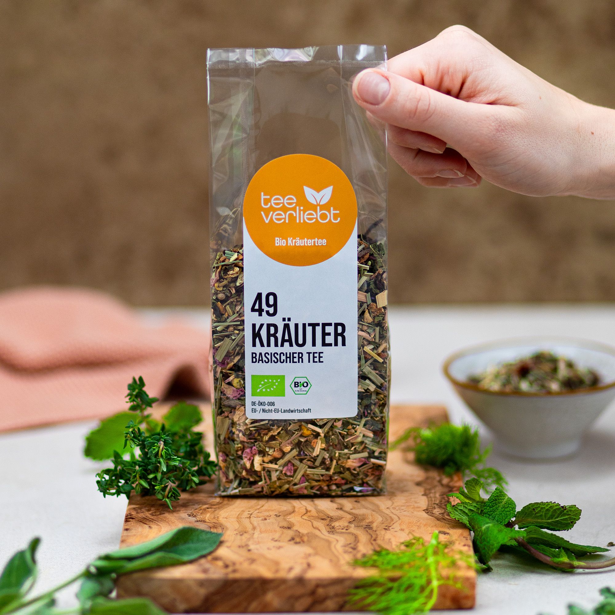 Hand hält transparente Verpackung mit Kräutermischung. Aufkleber mit Logo und Text: 49 Kräuter Basischer Tee Bio. Bio-Siegel.