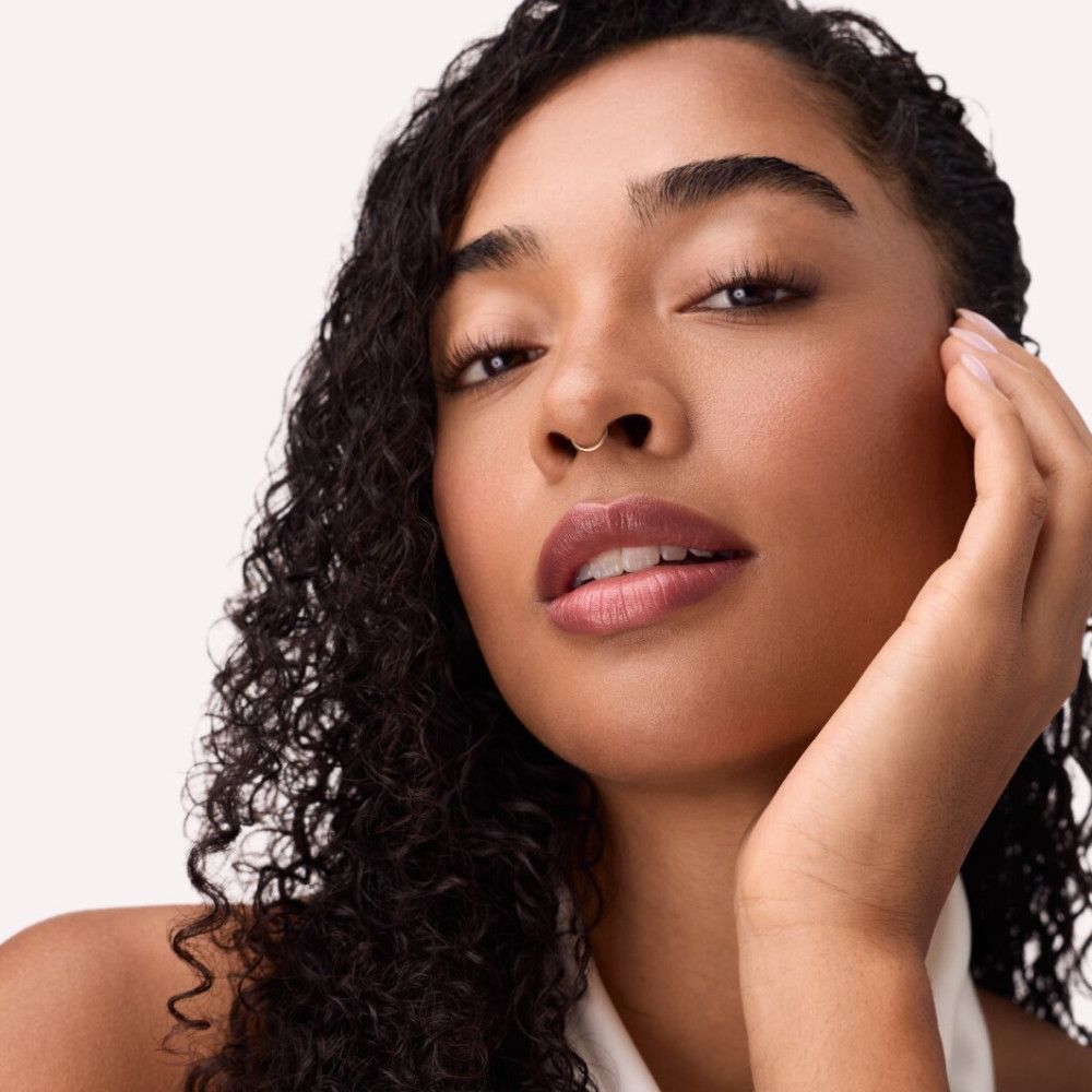 Porträt einer Frau mit rosafarbenem Gloss auf den Lippen. Sie berührt ihr Gesicht.