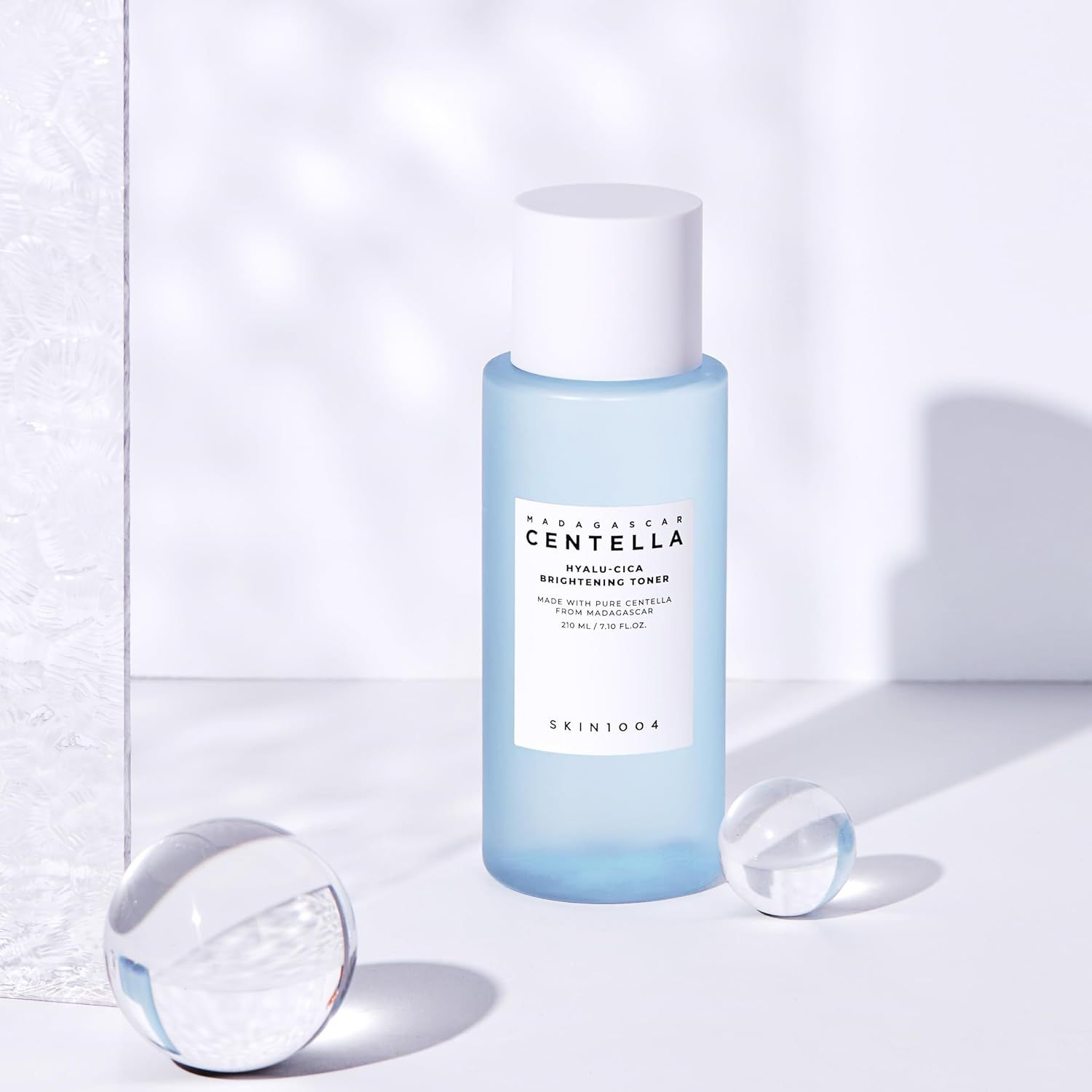 Hellblaue Flasche mit weißem Deckel. Aufschrift: MADAGASCAR CENTELLA HYALU-CICA BRIGHTENING TONER. Zwei Glaskugeln.
