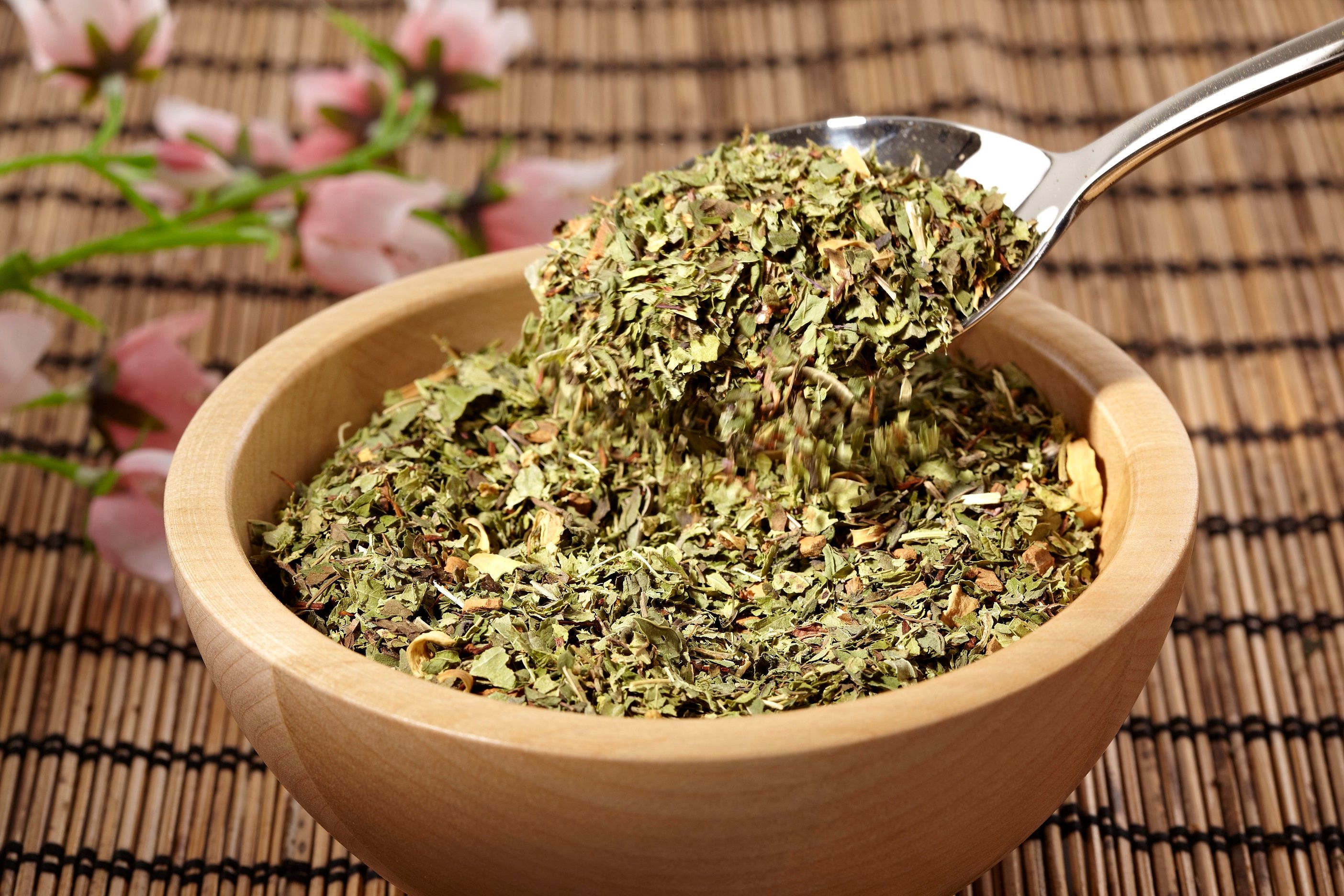 Holzschale mit Kräutermischung. Ein Löffel hebt Kräuter an. Hintergrund: Bambusmatte und Blüten.