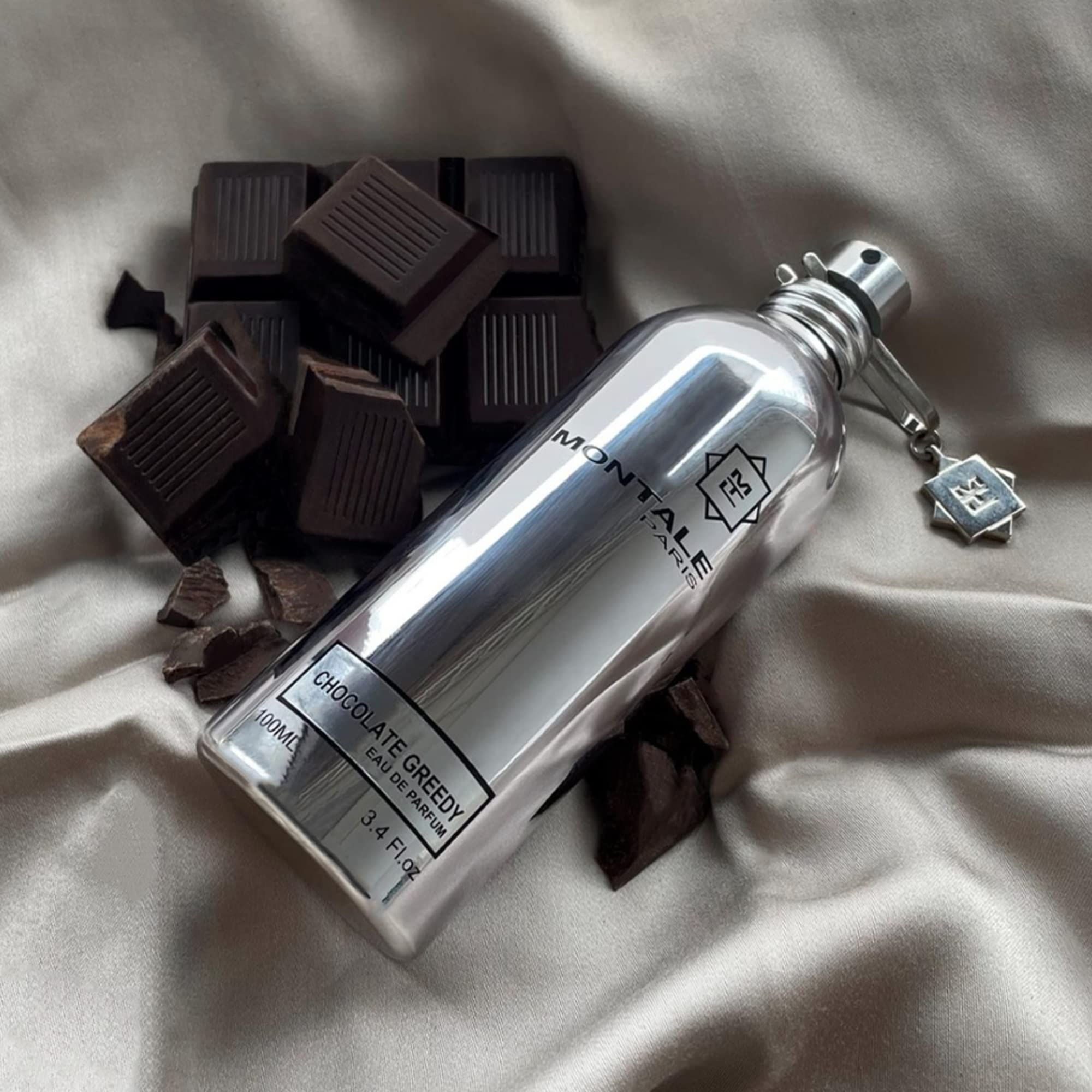 Silberne Parfumflasche mit Aufschrift Chocolate Greedy. Schokoladenstücke liegen daneben. Aufschrift Montale Paris.